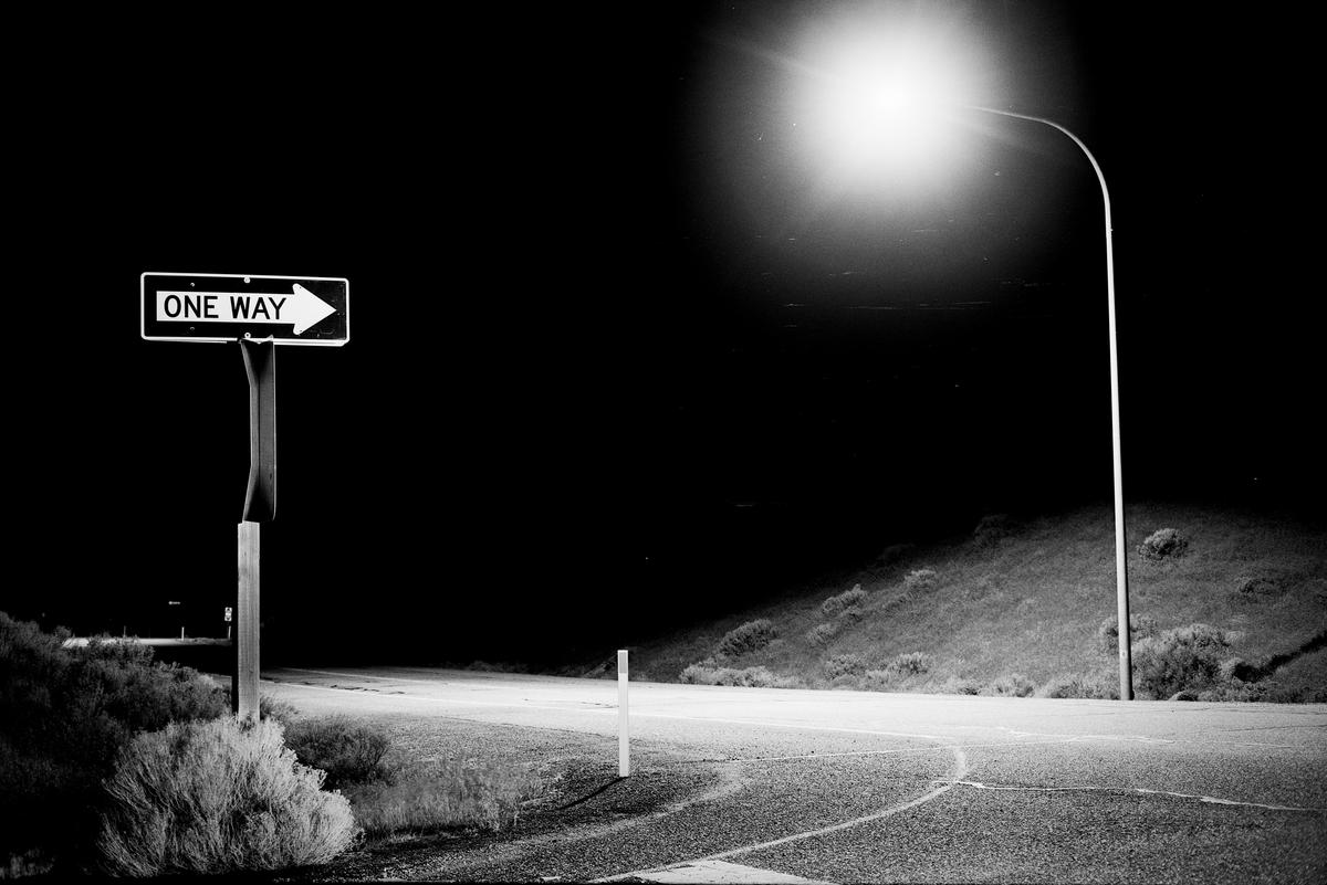 A one way sign stands next to a street light illuminating a road at night.