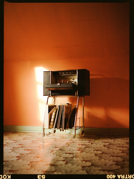 A record player on a cart with records sits in a room with an orange wall and patterned floor, illuminated by a shaft of sunlight.