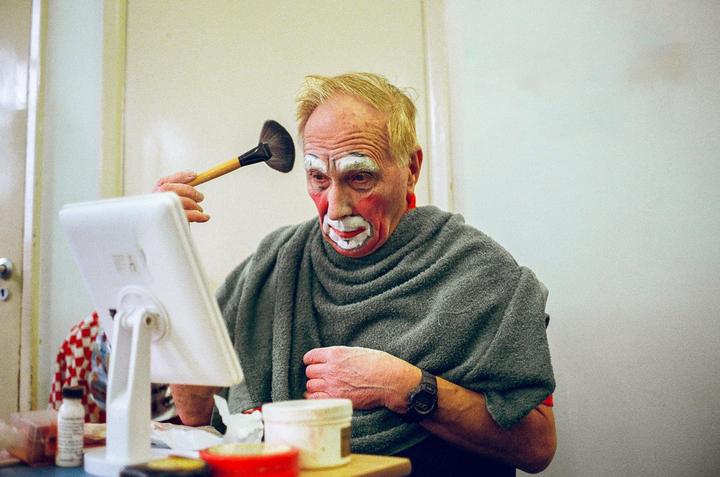 A man with clown makeup on his face applies more makeup with a brush while looking in a mirror.