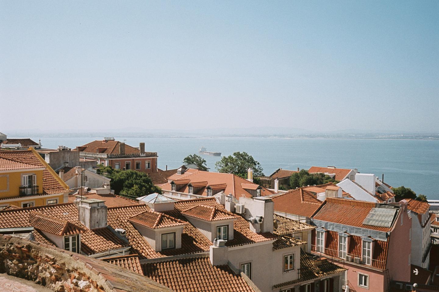 A cargo ship sails on the water behind a city with many buildings with red tile roofs.