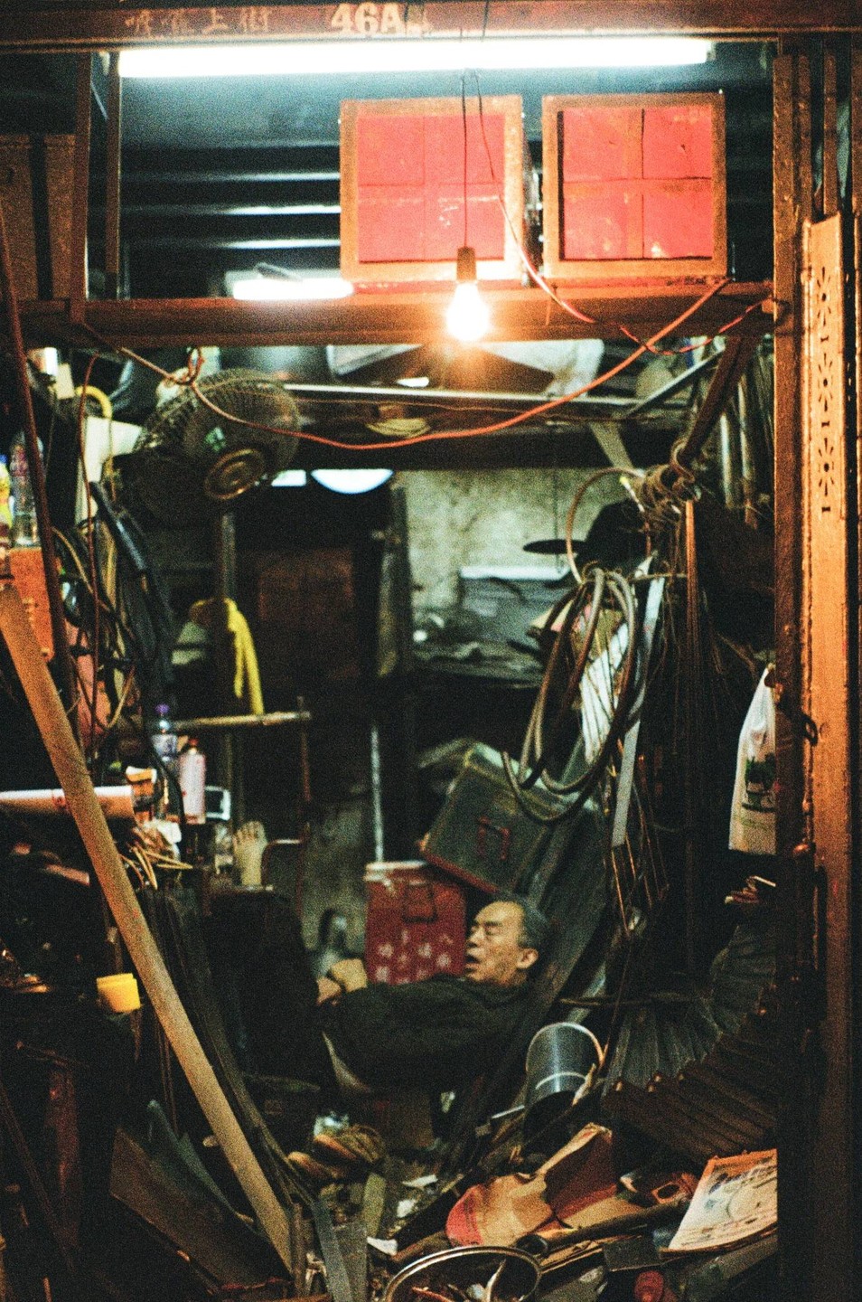 A man sleeps in a cluttered room with a single lightbulb illuminating the space.