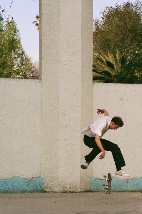 A man in a white shirt and black pants is captured mid-air while skateboarding next to a textured wall with a blue base.