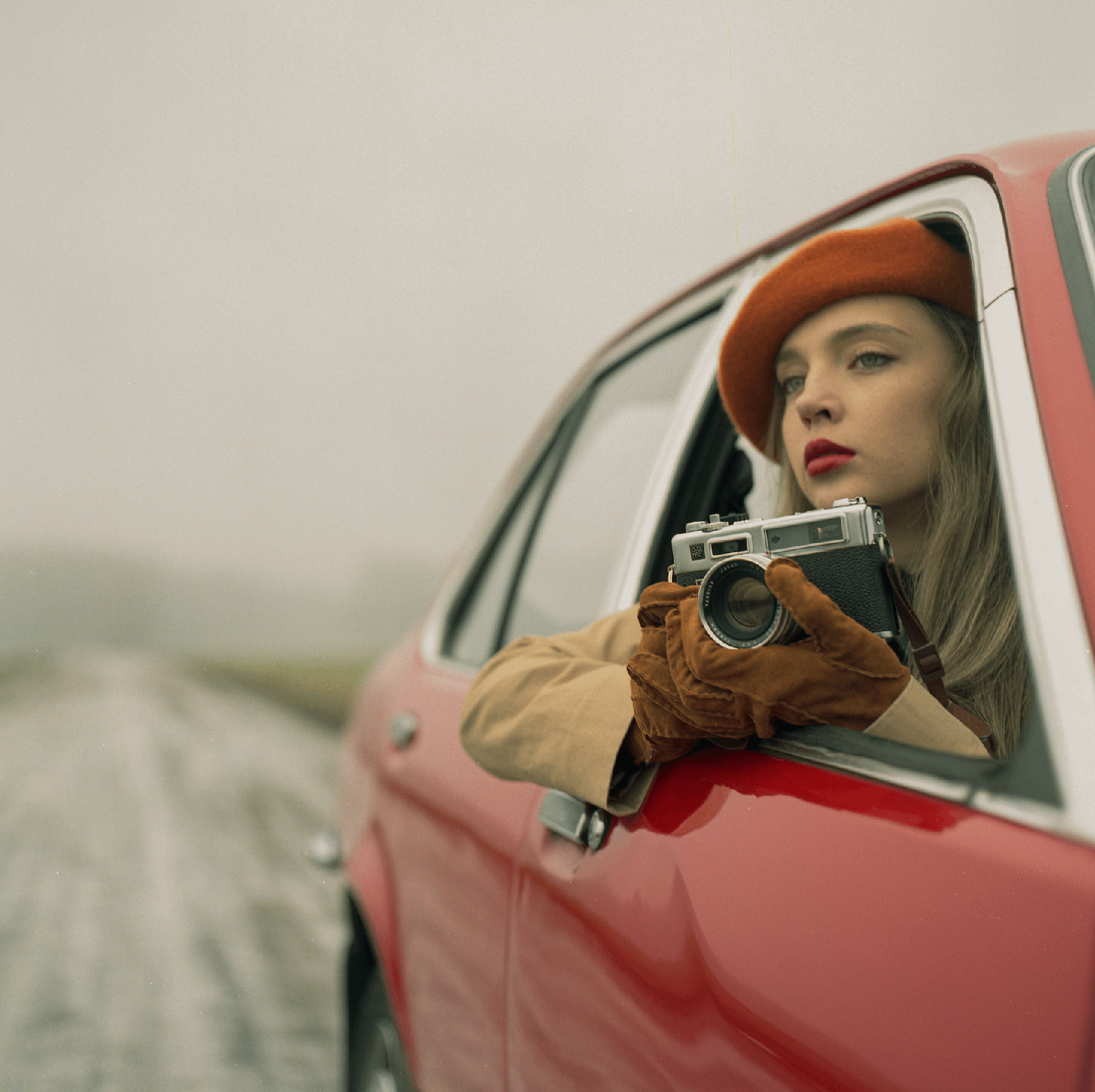 A woman in a red beret and brown gloves holds a camera out of the window of a red car.
