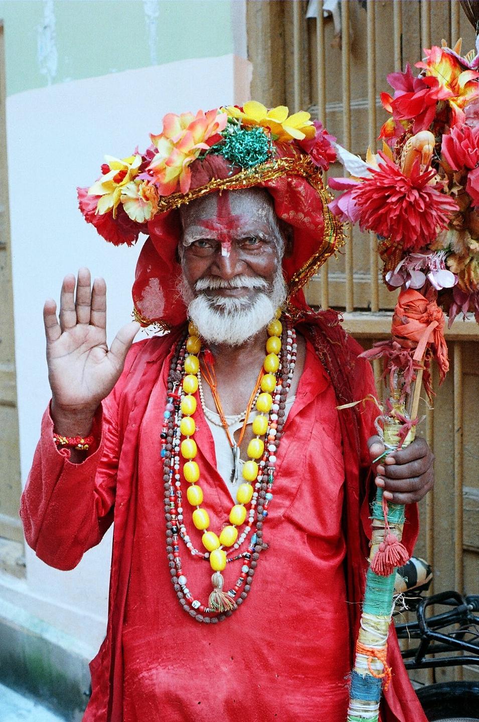 A man with a white beard and red markings on his face wears a red robe, colorful necklaces, and a floral hat while holding a decorated staff and raising his hand.
