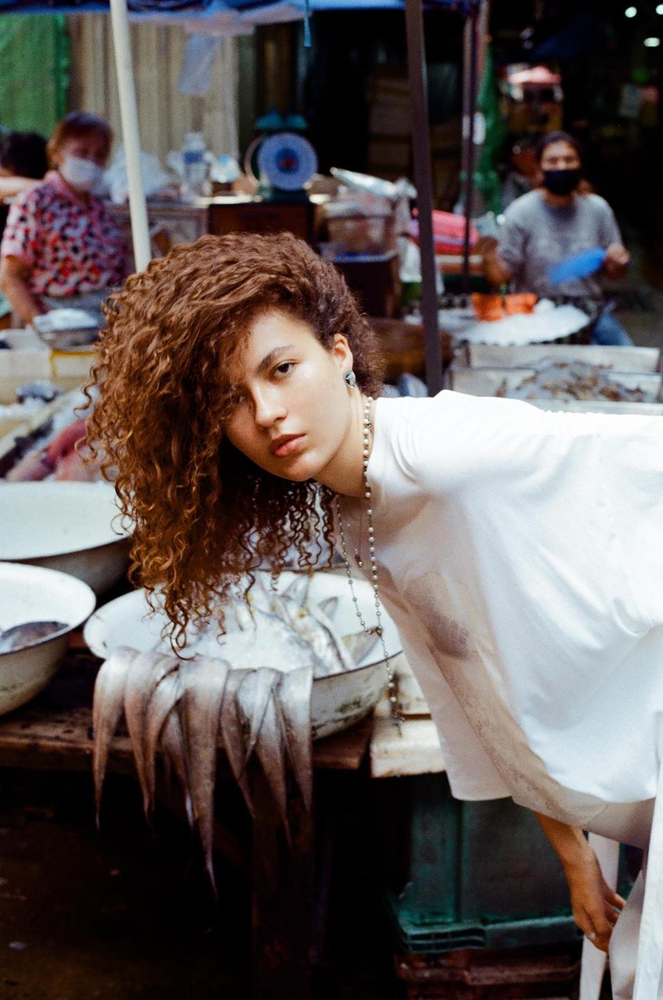 A woman with curly hair bends over a stall with fish in a market in Bangkok.