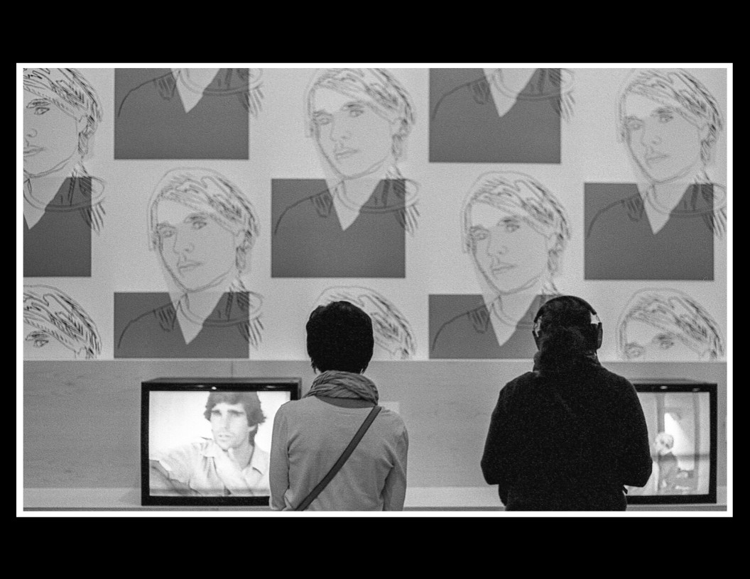 Two people stand with their backs to the viewer, looking at televisions and a wall with repeated images of a person's face.