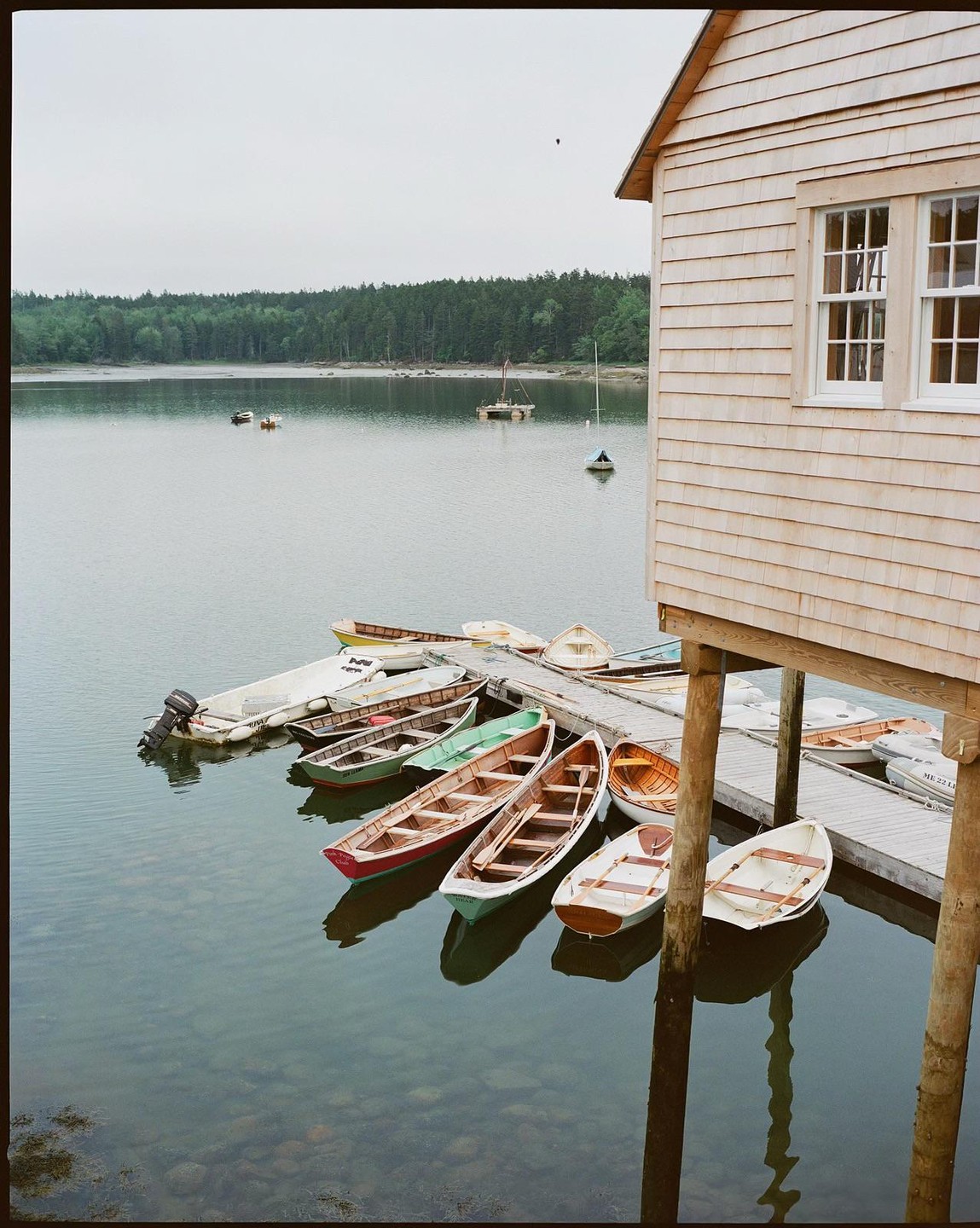 A wooden boathouse stands on stilts next to a dock with several small boats floating on calm water, with a forested shoreline in the background.