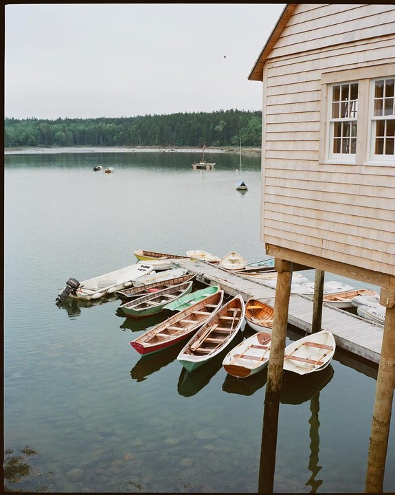 A wooden boathouse stands on stilts next to a dock with several small boats floating on calm water, with a forested shoreline in the background.