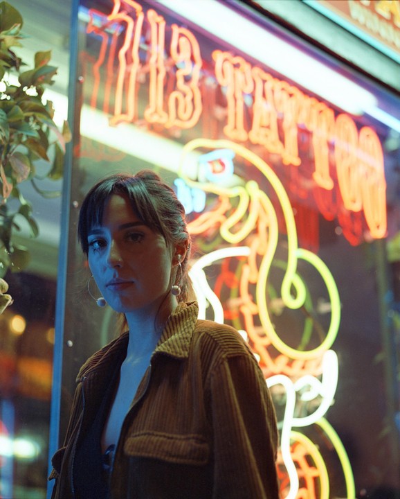 A woman stands in front of a neon sign for a tattoo parlor at night.