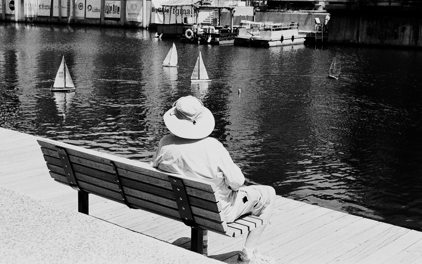 A person wearing a sun hat sits on a bench by the water, watching model sailboats.