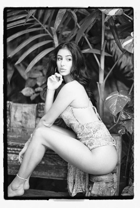 A woman sits on a wooden bench surrounded by large leafy plants.