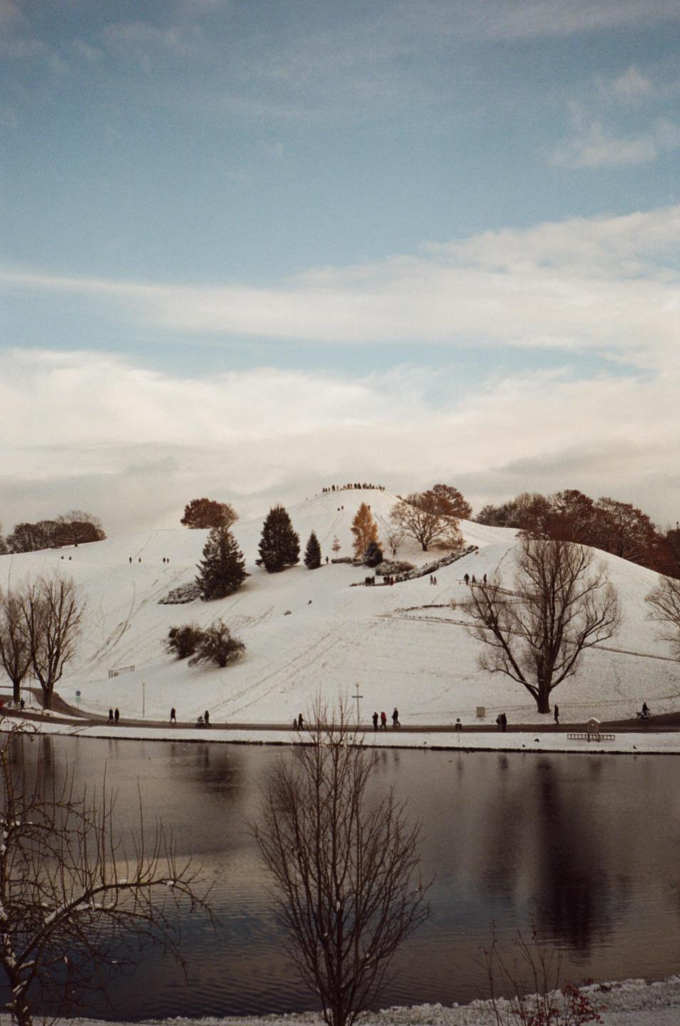 A snow-covered hill with trees and people walking on paths next to a lake under a cloudy sky.