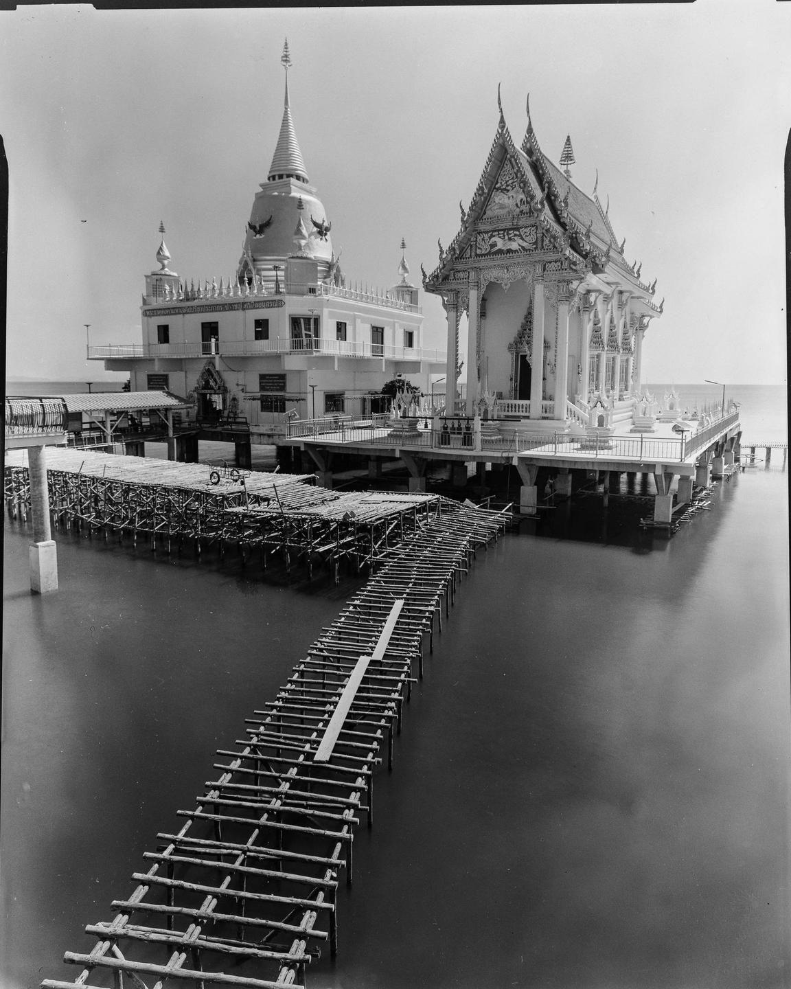 A temple and a stupa are built on stilts over water, connected by a wooden walkway under construction.