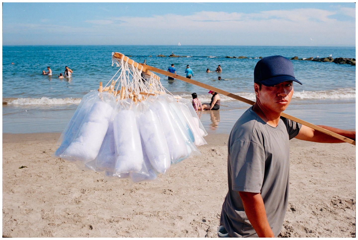 A man wearing a blue cap carries a pole with bags of cotton candy on a beach with people swimming in the ocean.