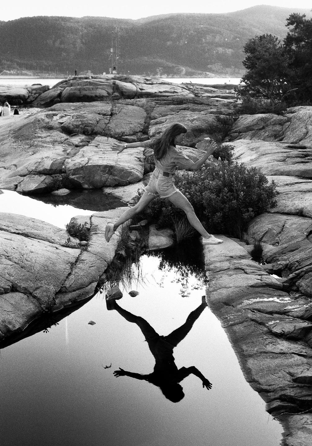 A woman jumps over a puddle on a rocky shore, her reflection visible in the water.