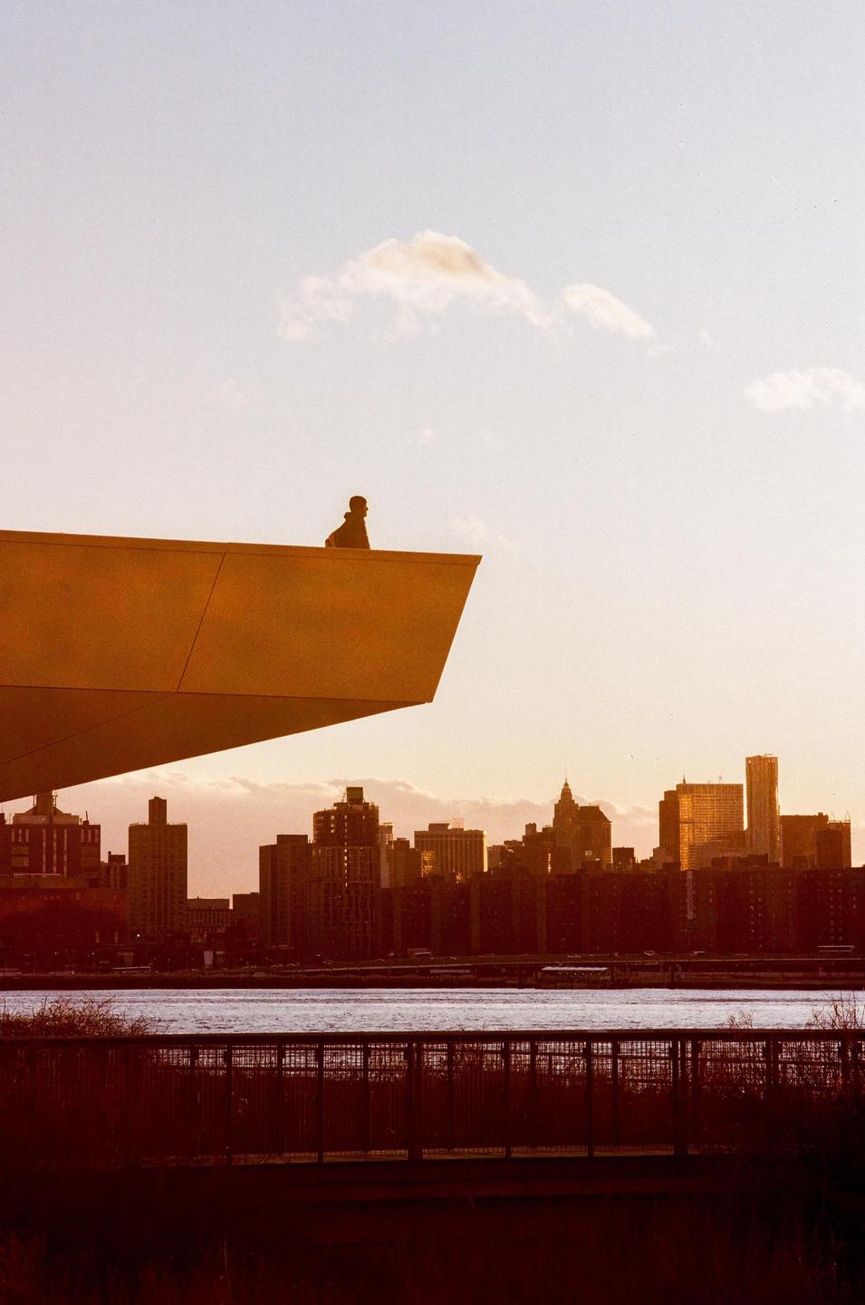 A person sits on the edge of a modern building overlooking a city skyline and river at golden hour.
