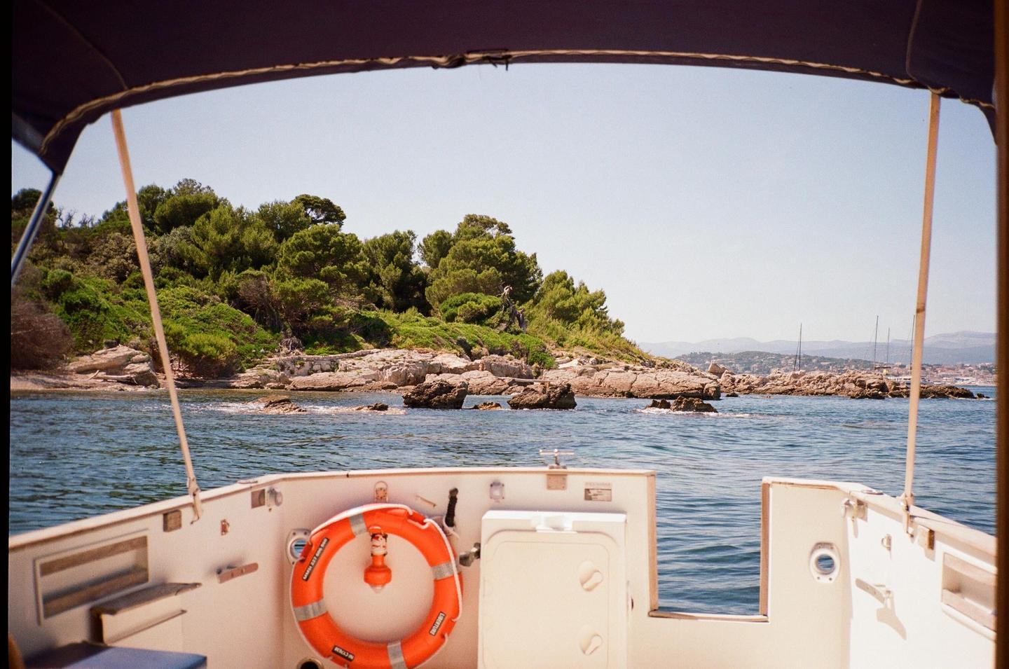 A view from a boat shows a rocky coastline with green trees and distant mountains under a clear sky.