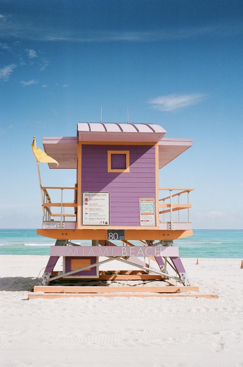 A purple and orange lifeguard stand with a yellow flag stands on a white sand beach under a clear blue sky.