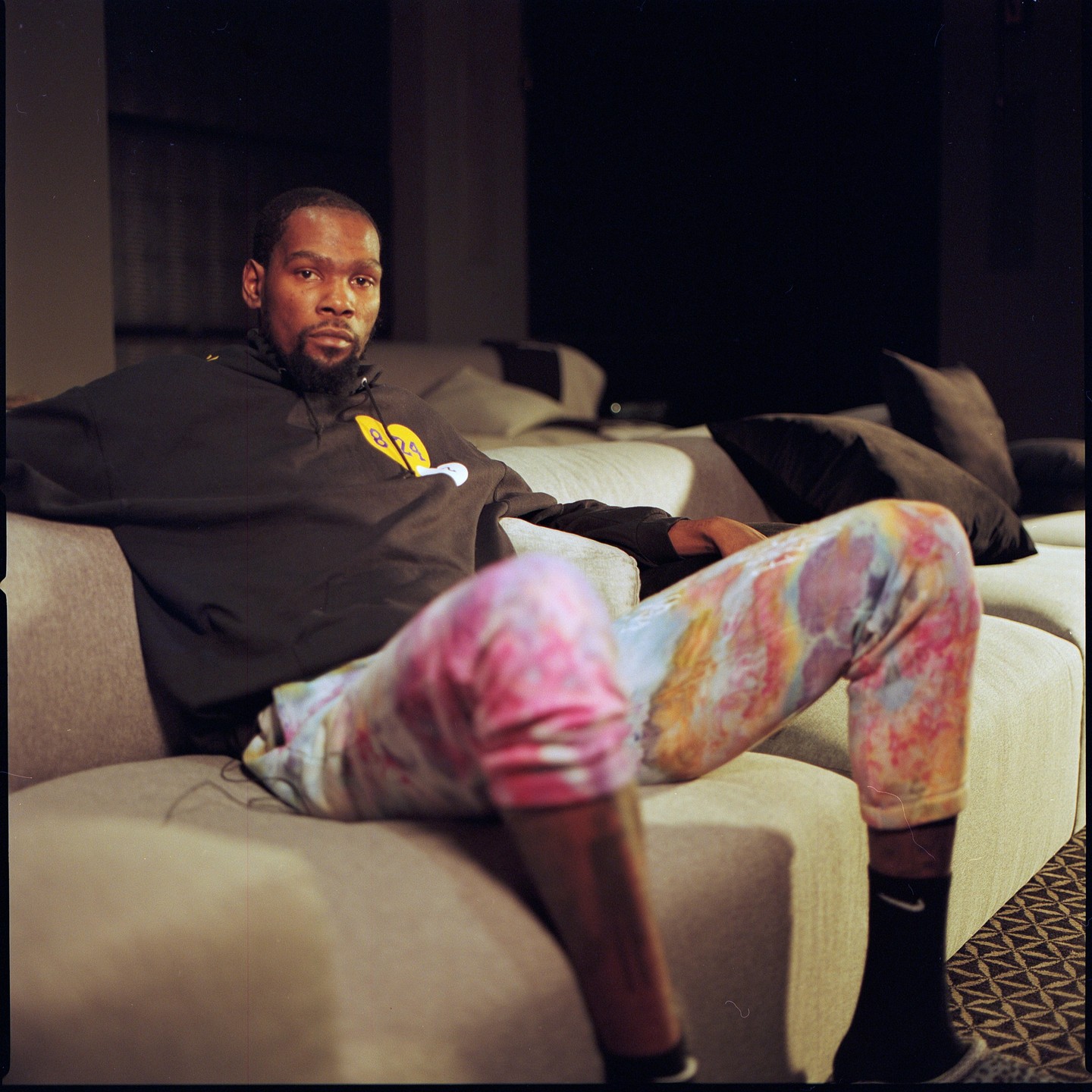A man with a beard is sitting on a sofa, wearing a black hoodie and colorful pants.