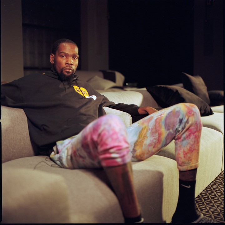A man with a beard is sitting on a sofa, wearing a black hoodie and colorful pants.