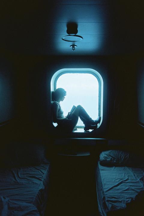 A woman sits in a window reading a book in a dark room with two beds.