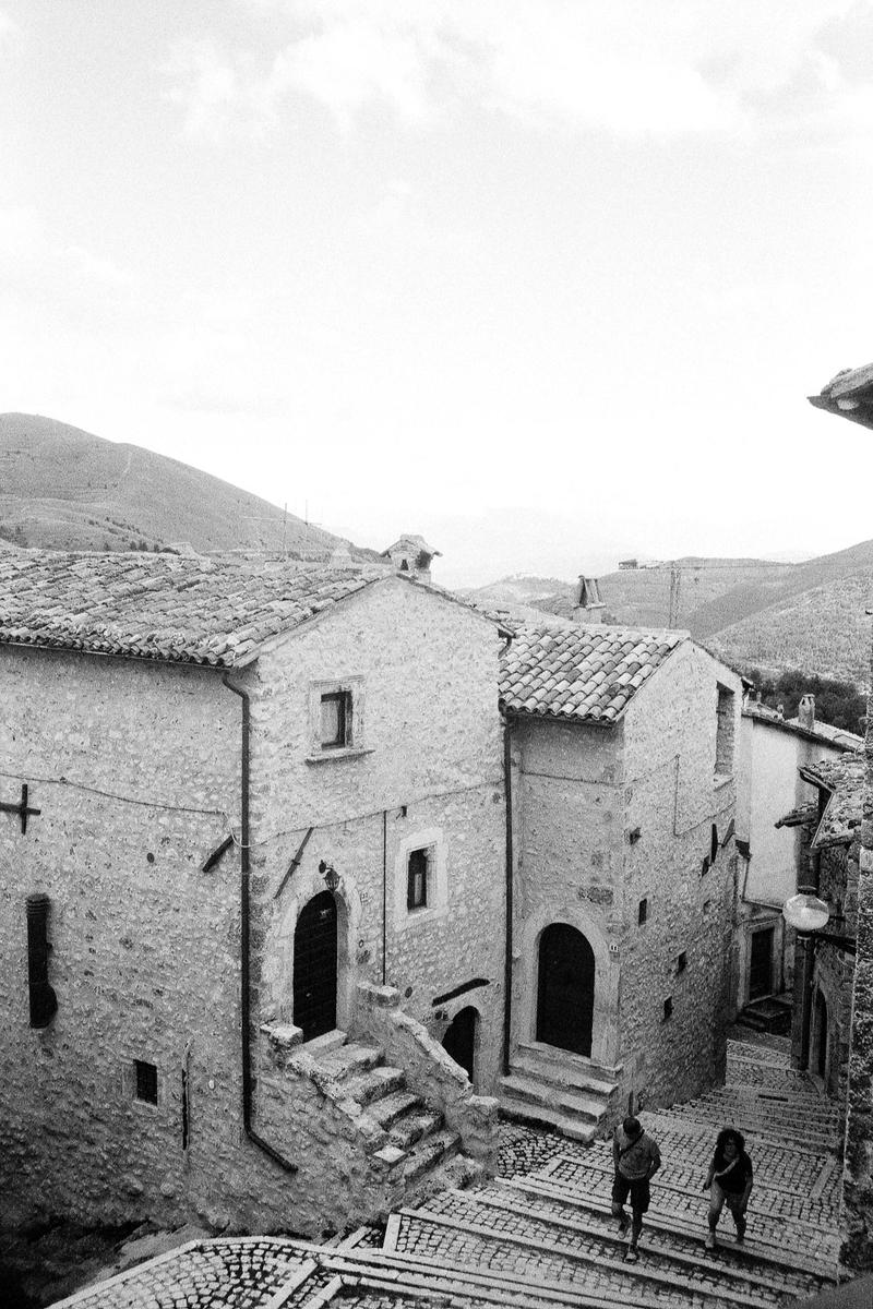 Two people walk down a cobblestone street between stone buildings in a mountain village.