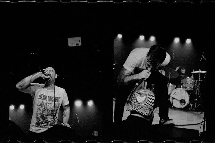 Two men perform on stage with a drummer in the background, illuminated by spotlights.