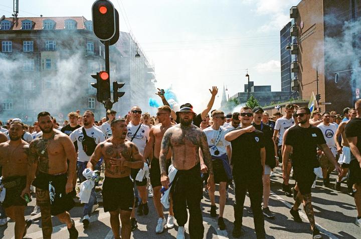 A large group of men march down a city street, some shirtless and some holding blue smoke flares, with buildings and traffic lights in the background.