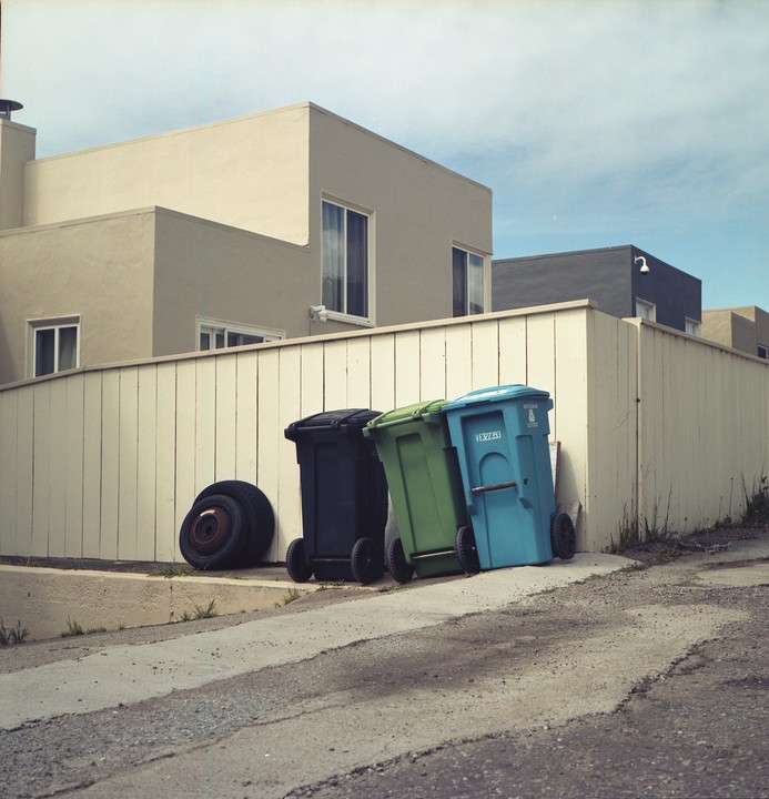 Three trash cans and two tires are lined up against a fence in front of a building.