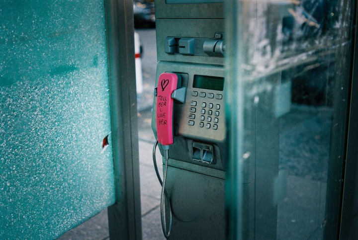 A pink telephone receiver with a message written on it hangs in a public telephone booth.