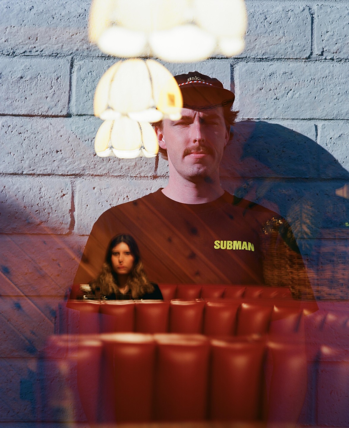 A man and a woman are visible through a double exposure, with the man in the foreground against a brick wall and the woman in the background sitting in a red booth.
