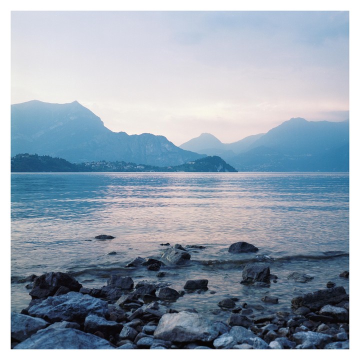 A body of water with rocks in the foreground is surrounded by mountains under a pale sky.