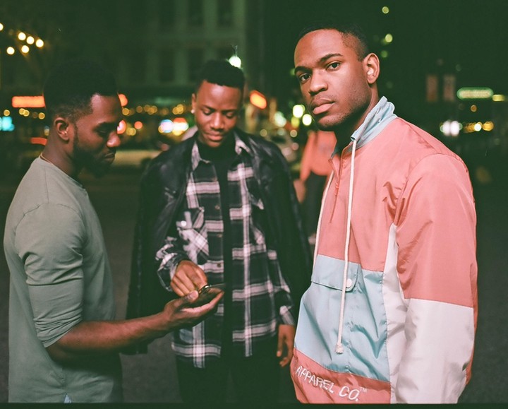 Three men are standing on a street at night, with one man looking at a phone held by another, and the third man looking directly at the viewer.
