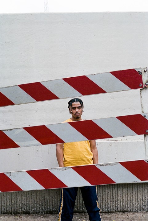 A man with braided hair stands behind a red and white striped barrier.