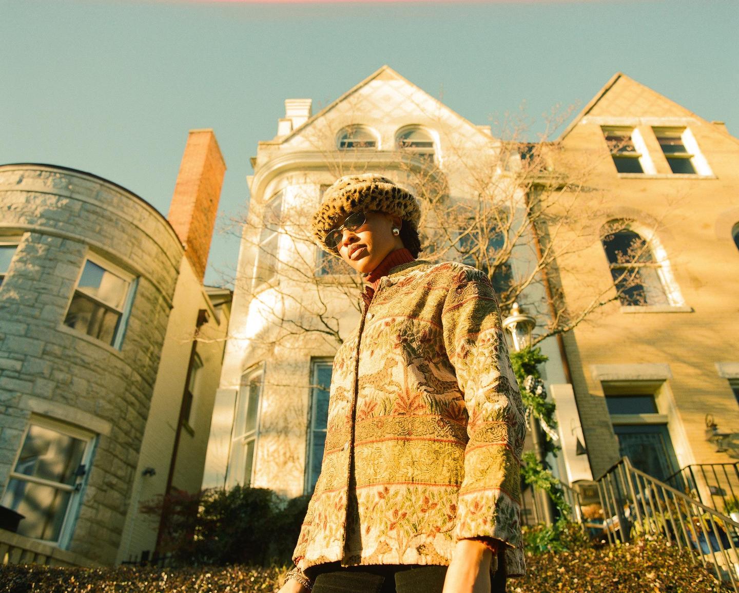 A woman wearing a patterned jacket and a leopard print hat stands in front of a large house.