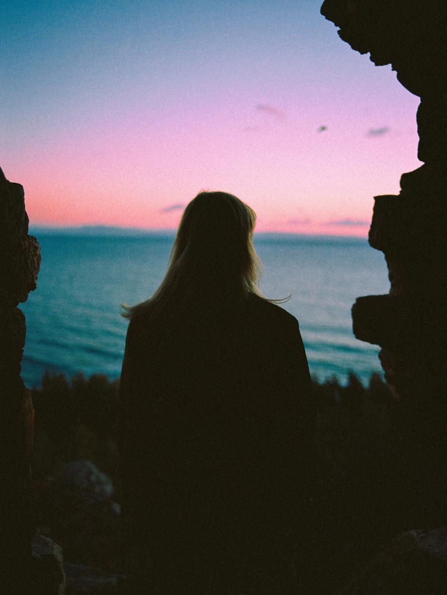 A woman looks out at the ocean during sunset, framed by dark rock formations.
