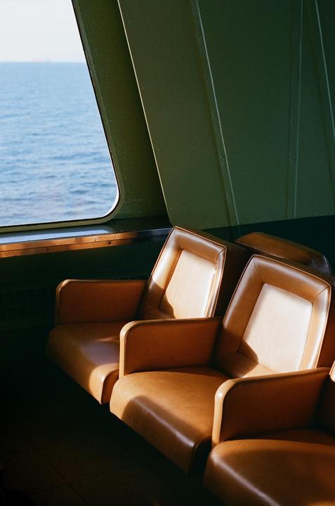 Sunlight illuminates a row of empty brown seats next to a window overlooking the ocean on board the M.V. Coho.