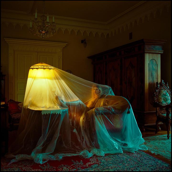 A woman is partially visible behind a plastic sheet draped over a lamp and furniture in a room with ornate decor.
