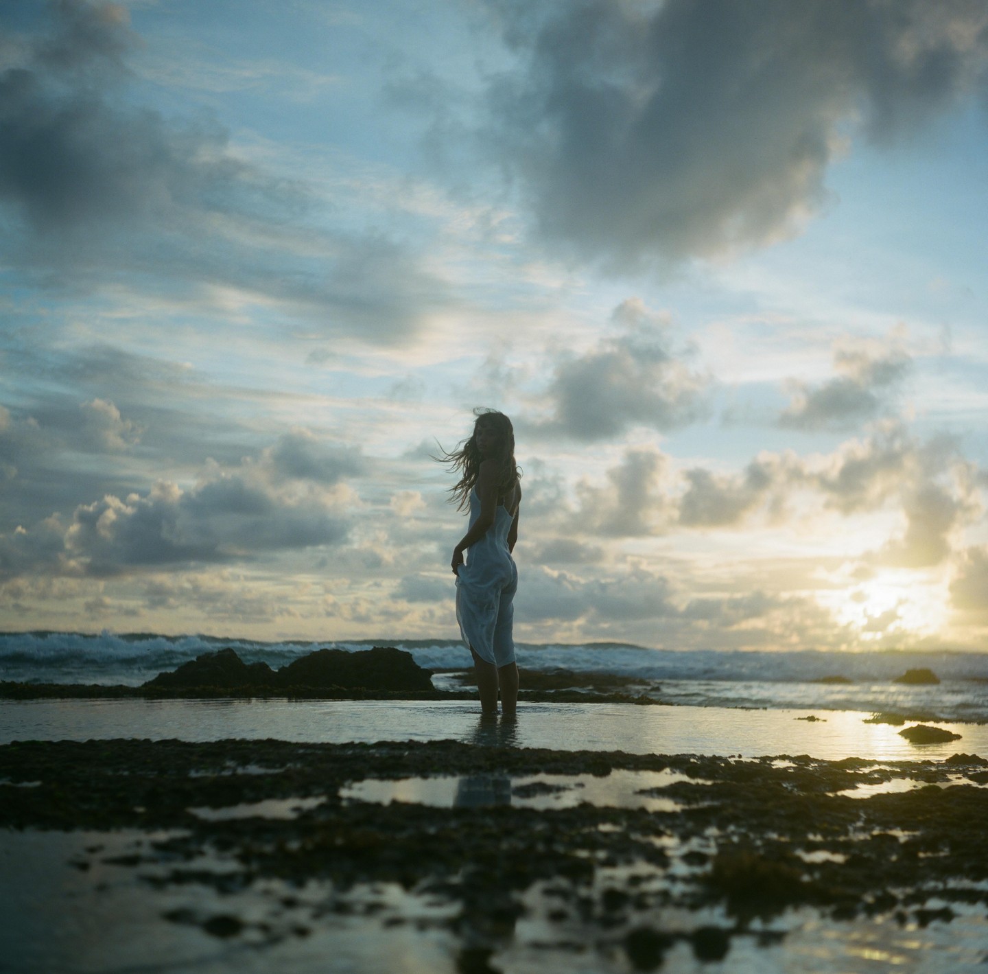 A woman stands in shallow water on a rocky beach with the sun setting over the ocean.