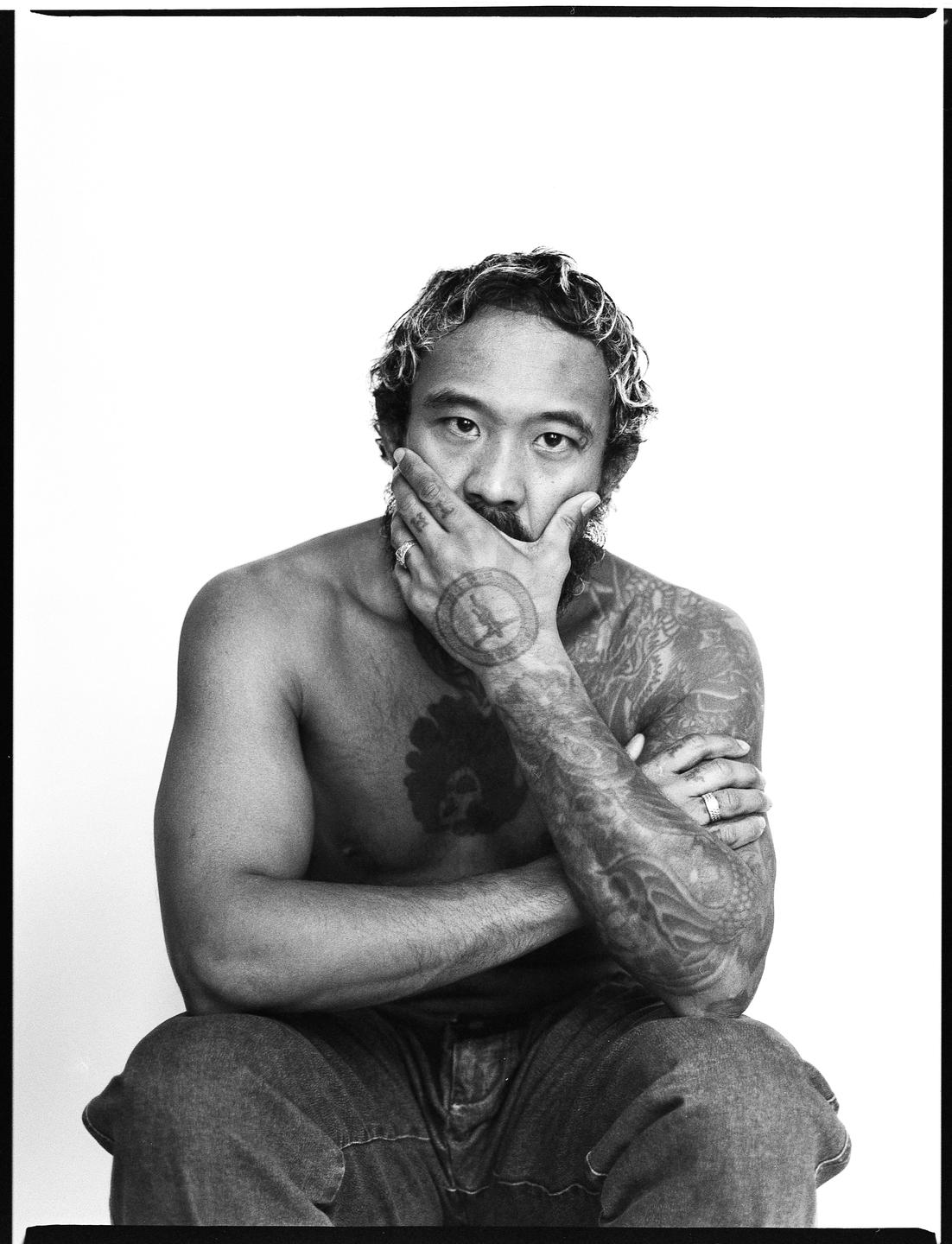 A man with tattoos on his chest and arms sits with his hand covering his mouth.