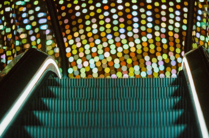 An escalator ascends towards a wall of colorful, out-of-focus lights.