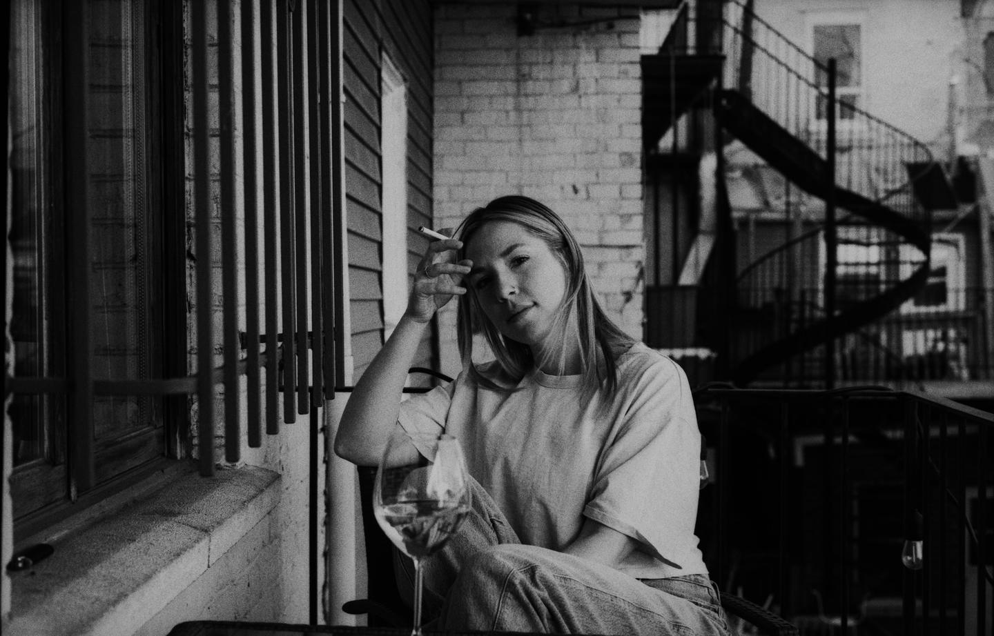 A woman sits on a balcony with a cigarette and a glass of wine.