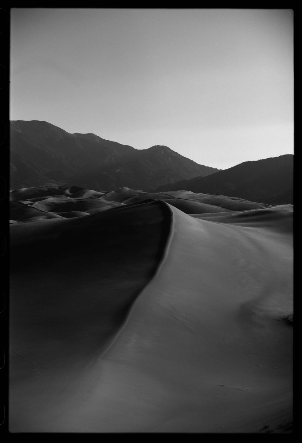 A black and white landscape shows sand dunes with mountains in the background under a clear sky.