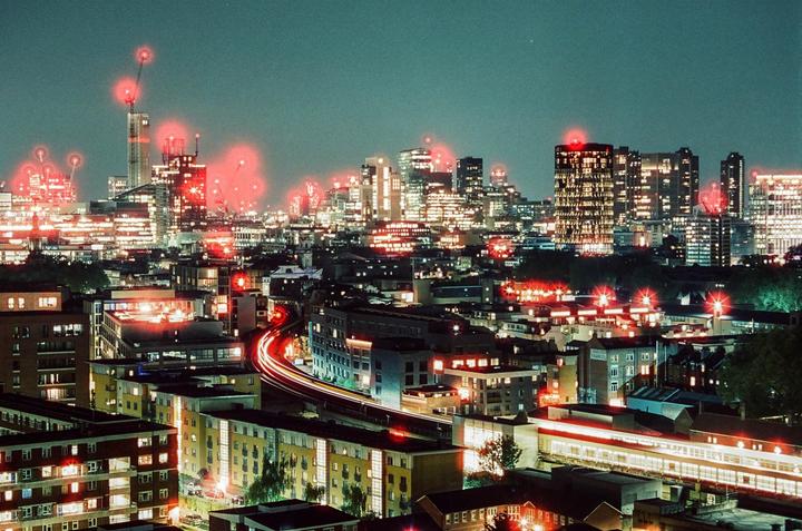 A cityscape at night shows buildings with illuminated windows and red lights on top of skyscrapers, with light trails from moving vehicles in the foreground.