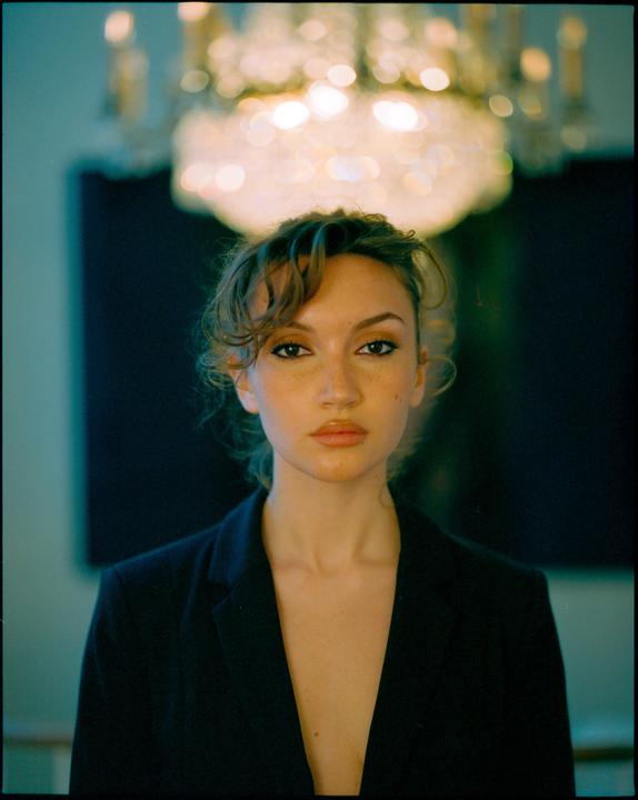 A woman with dark hair and freckles looks directly at the viewer, with a chandelier visible above her head.