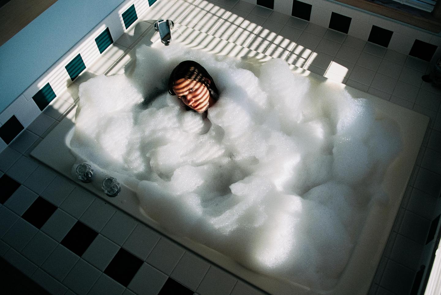 A woman is taking a bubble bath in a tiled bathroom with strong shadows from a window blind.