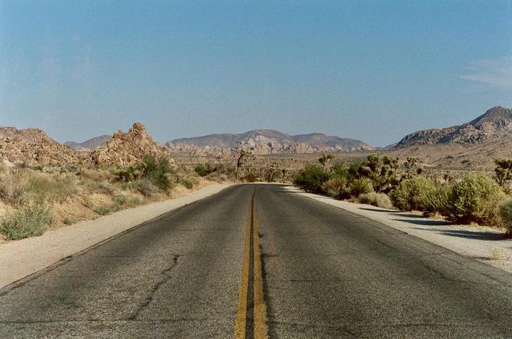 A long, straight road with a double yellow line stretches through a desert landscape with mountains and sparse vegetation under a clear sky.