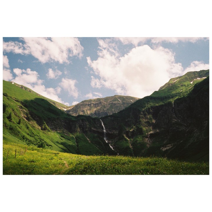 A waterfall cascades down a lush green mountain under a partly cloudy sky.