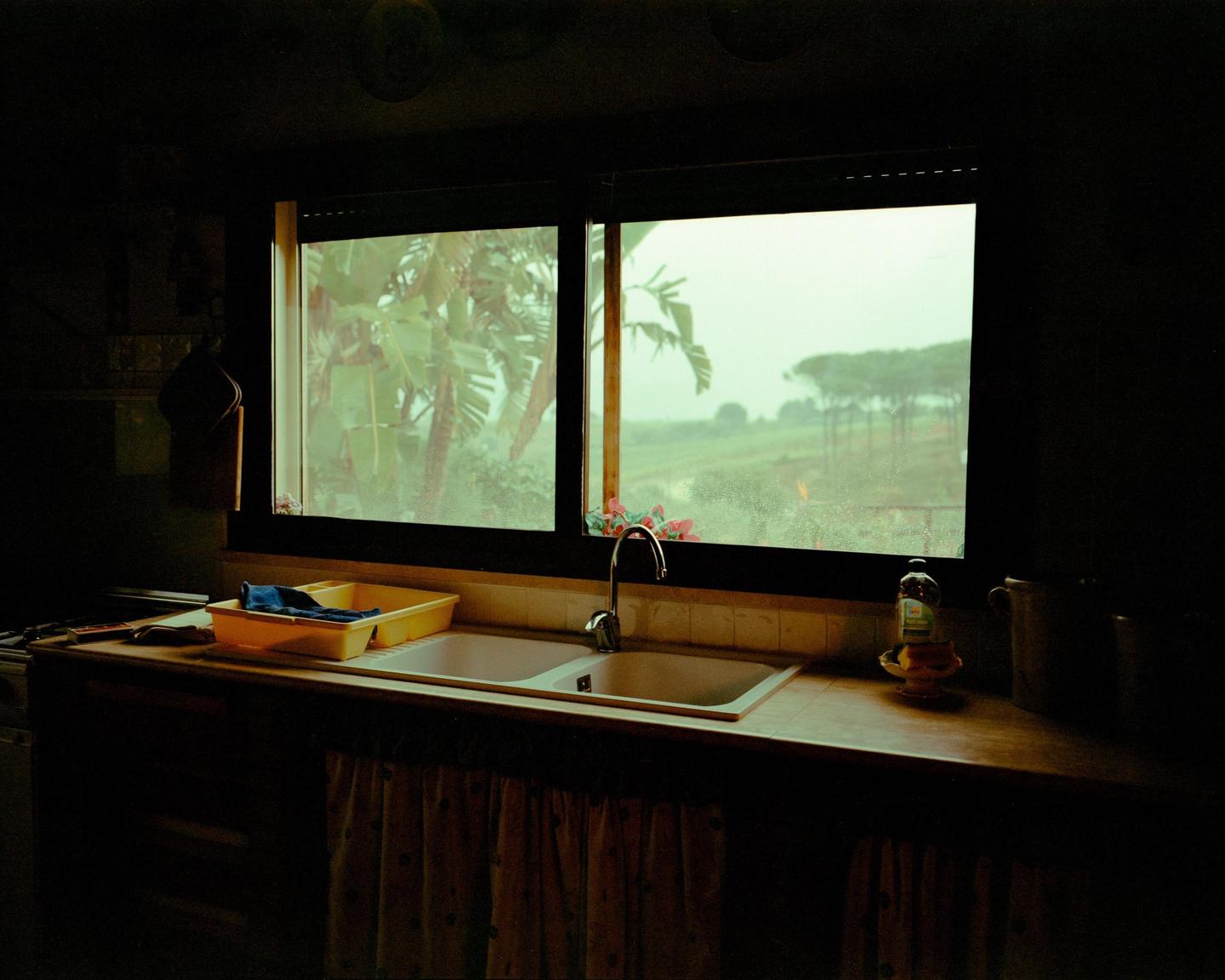 A kitchen sink and counter are visible in the foreground, with a window looking out onto a green landscape with trees in the background.