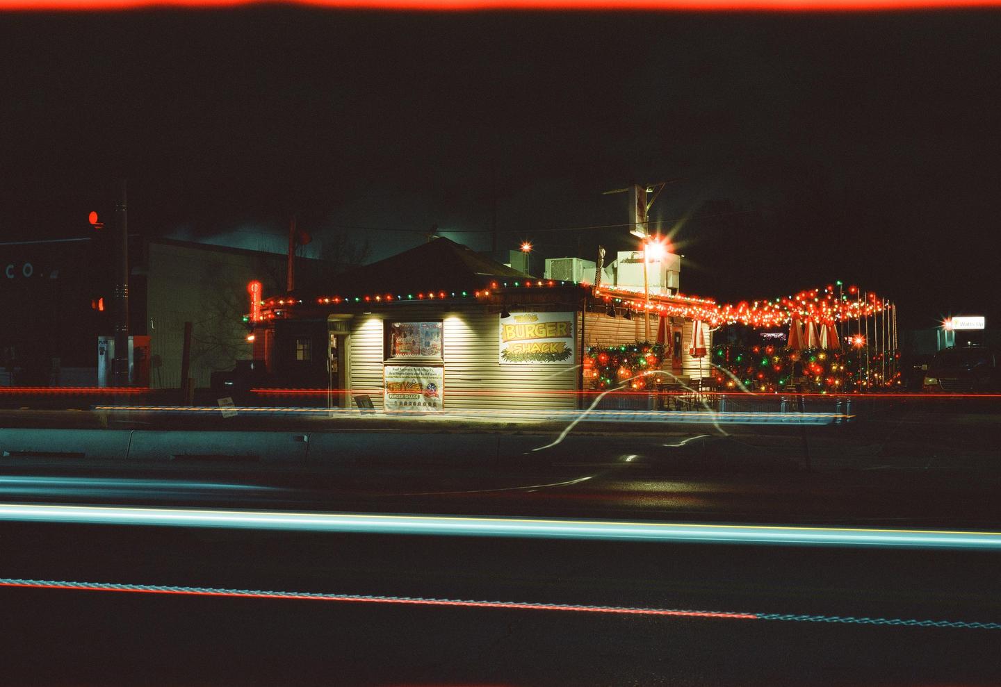 A burger shack decorated with Christmas lights is illuminated at night with light trails from passing vehicles in the foreground.